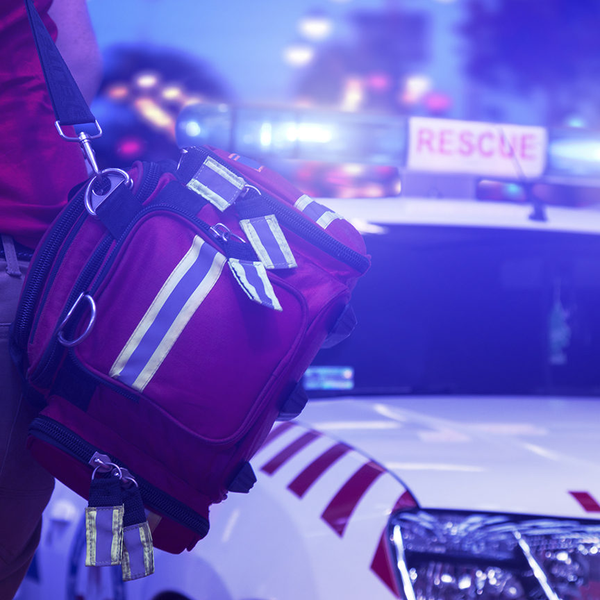 Back view of rescue service team member holding a bag and standing next to the vehicle with rotating beacons